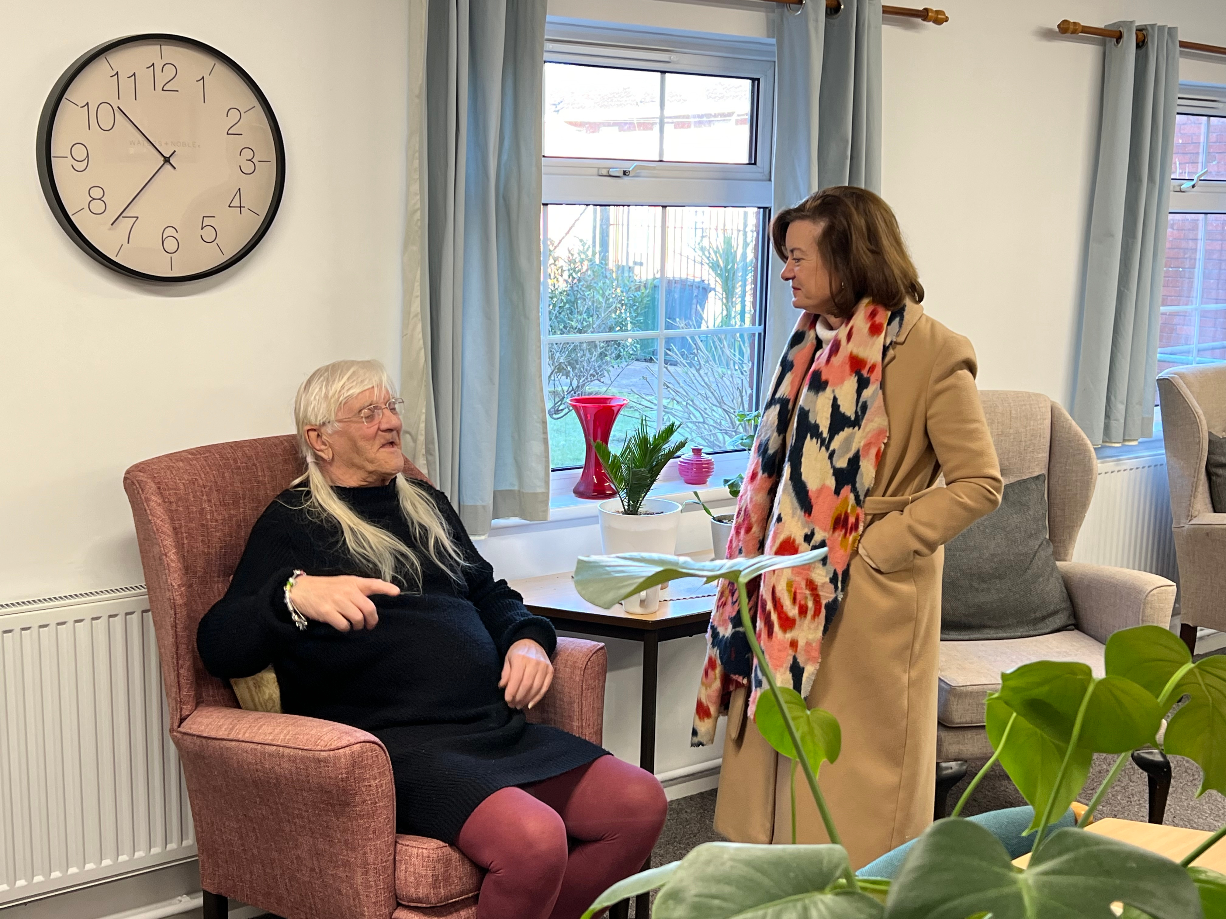 First Minister visiting a housing resident. Eluned Morgan is standing next to an elderly resident in an armchair mid coversation with nutural expressions.