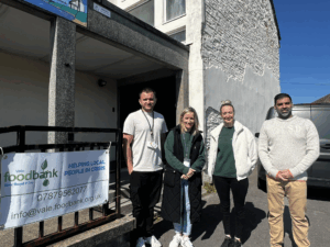 Four people smiling next to a banner that reads Vale Foodbank, Helping local people in crisis