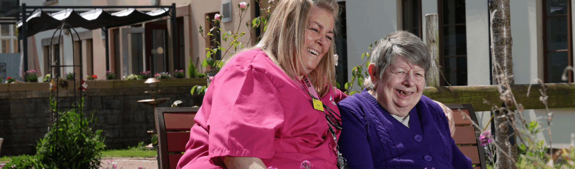 Female care home resident sitting on bench in garden with carer smiling